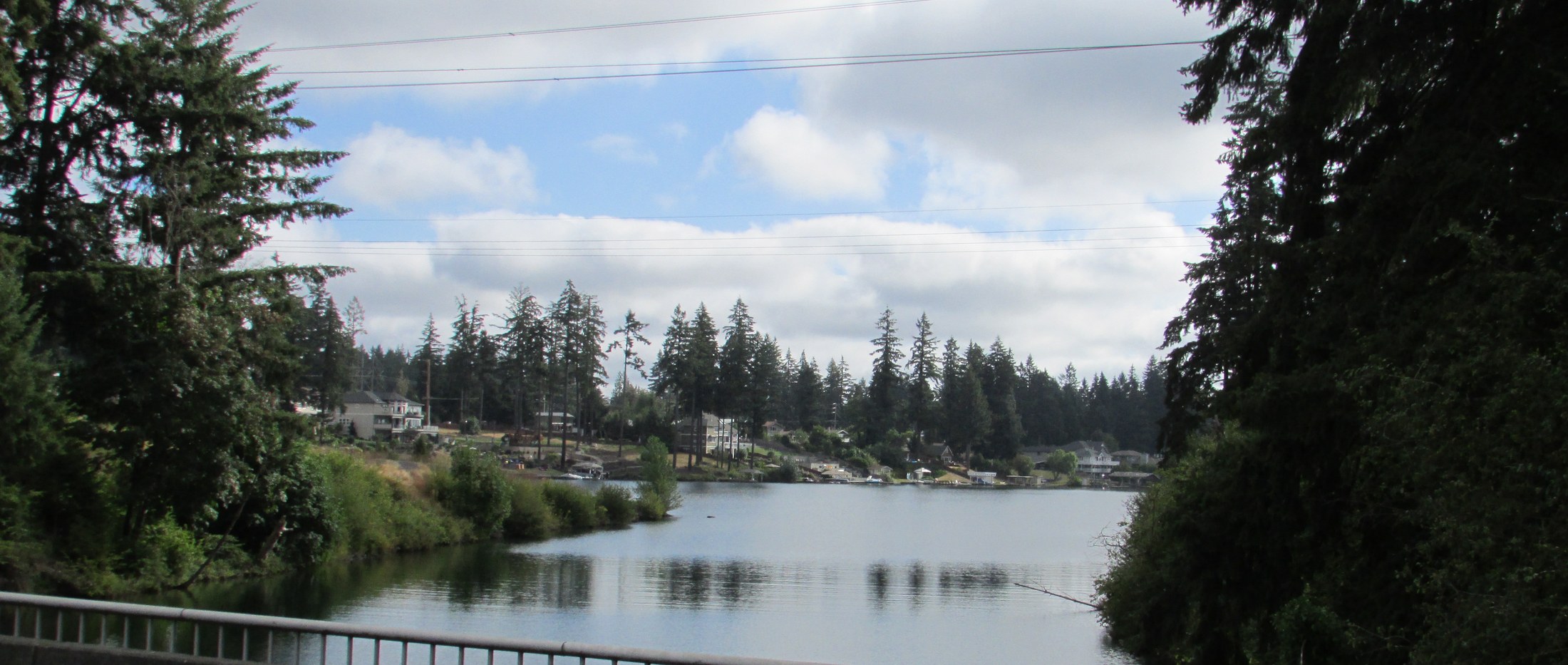 Lake Tapps at the outlet basin, calm water with a forested shoreline and low hills in the distance under an overcast sky